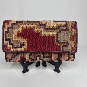 May include: A rectangular clutch with a geometric pattern in shades of red, brown, and tan. The clutch is displayed on a black stand against a white background. The flap closure is a darker shade of red.
