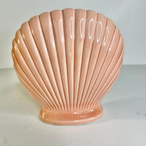 Könnte beinhalten: Ein dekoratives Objekt in Form einer Muschelschale aus Keramik in einem sanften, gedämpften Rosa. Die Schale hat eine gerippte Textur mit glänzendem Finish. Der Sockel hat eine abgerundete Form, und das Gesamtdesign erinnert an Küstendekoration.