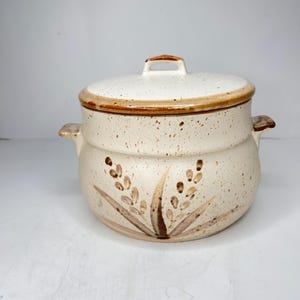 May include: Cream-colored ceramic casserole dish with lid. The dish features a speckled finish and brown floral design. The lid has a handle and a brown rim. The casserole dish has two side handles.