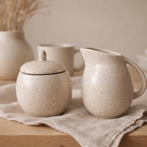 Könnte beinhalten: Ein cremefarbener Keramik-Zuckertopf mit Deckel und ein kleiner Sahnekrug mit gesprenkeltem Design. Im Hintergrund eine Vase mit Trockenblumen und eine Tasse, alles auf einer Holzoberfläche.