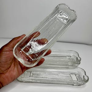 May include: Clear glass corn-shaped serving dishes. The dishes have a textured surface and a rectangular shape with a curved handle. Three dishes are visible, with one held in a hand.
