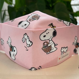 May include: Pink fabric face mask with a white Snoopy character carrying a basket of flowers. The mask has a white heart with the text "Athleticable" on it. The mask is labeled with the number "#1".