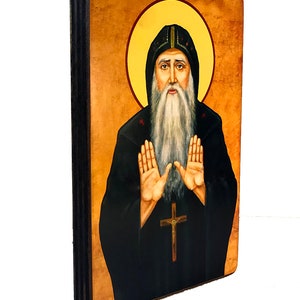 Op de afbeelding: Een handgeschilderd houten icoon van een man met een baard in een zwarte robe met een halo. Hij houdt zijn handen omhoog in een gebaar van gebed. Het icoon is op een houten achtergrond.