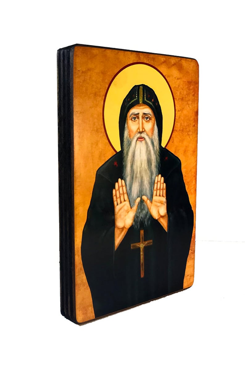 Op de afbeelding: Een handgeschilderd houten icoon van een man met een baard in een zwarte robe met een halo. Hij houdt zijn handen omhoog in een gebaar van gebed. Het icoon is op een houten achtergrond.