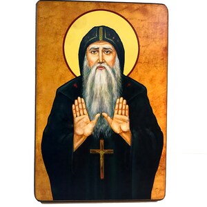 Op de afbeelding: Een religieus icoon van een man met een baard en een halo, die een zwarte robe en een kruis draagt. Het icoon is geschilderd op een houten paneel met een gouden achtergrond.