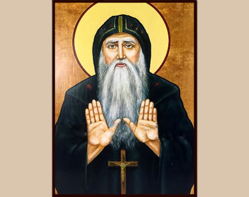 Op de afbeelding: Een religieus icoon schilderij van een man met een lange witte baard en een zwarte robe. Hij heeft een halo boven zijn hoofd en houdt zijn handen omhoog in een gebaar van gebed. De achtergrond is goudkleurig.