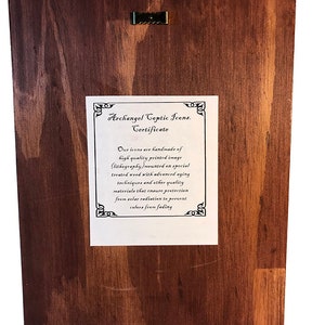 May include: A certificate for an Archangel Coptic Icon. The certificate is printed on white paper and mounted on a dark brown wooden background. The certificate states that the icons are handmade of high quality printed image (lithography) mounted on special treated wood with advanced aging techniques and other quality materials that ensure protection from solar radiation to prevent colors from fading.
