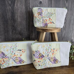 May include: Three quilted zipper pouches featuring a patchwork star design in shades of yellow, purple, orange, and white. Each pouch has a white zipper closure and a light green and white checkered fabric accent on the side of the zipper.