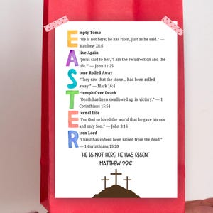 Peut inclure: Un sac cadeau rouge avec une carte blanche avec le mot « EASTER » en lettres colorées. La carte comprend des versets bibliques sur la résurrection, avec un dessin de trois croix sur une colline.