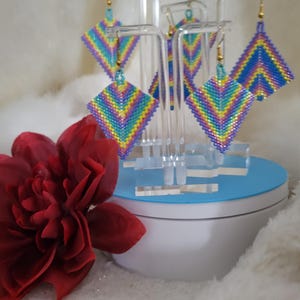 May include: Beaded square earrings in a rainbow chevron pattern. The earrings are displayed on clear stands with gold-colored hooks. A large, red fabric flower is in the foreground. The earrings are a vibrant mix of colors.
