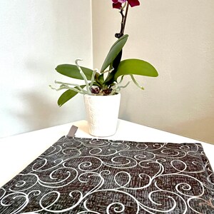 May include: A potted orchid plant with a single purple flower in a white pot. The plant is sitting on a brown and white patterned table runner.