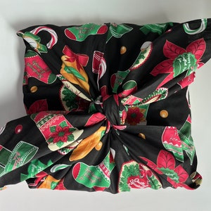 May include: A black fabric wrapping a square object, patterned with Christmas stockings, poinsettias, and candy canes in red, green, and white. The fabric is tied in a knot, creating a decorative presentation.
