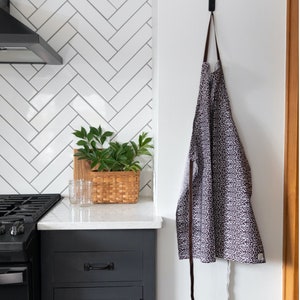 Puede incluir: Un delantal de cocina con estampado blanco y negro colgado de un gancho en una pared blanca. El delantal tiene correas marrones y está hecho de tela.