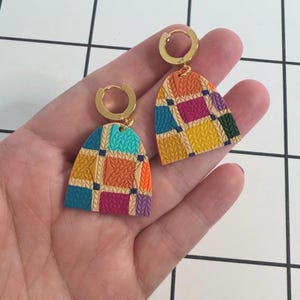 May include: A pair of dangle earrings featuring gold-toned circular hoops attached to arch-shaped pendants. The pendants display a multi-colored patchwork design with a textured, knitted-style pattern in shades of teal, yellow, orange, and magenta.