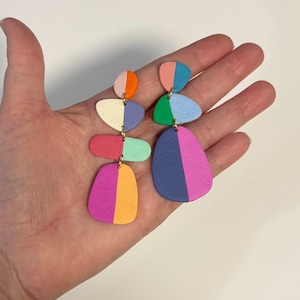 May include: A pair of mismatched dangle earrings featuring stacked, pebble-shaped components with a textured matte finish. The earrings display a vibrant color palette including orange, cream, purple, red, mint green, pink, blue, and dark blue.