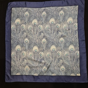 May include: A square silk scarf featuring a repeating peacock feather pattern in shades of blue, grey, and white. The central design is framed by a solid blue border. A small white label with black text is visible in the bottom right corner.