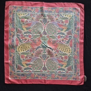 May include: A square silk scarf featuring a symmetrical pattern of four peacocks in shades of green, pink, and gold, surrounded by a floral border and a solid dusty rose frame. The word LIBERTY is printed in the bottom right corner of the design.