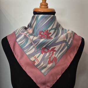 May include: A square silk scarf with a floral design. The scarf features a pink border and a central pattern of red flowers and diagonal stripes in shades of gray, blue, and white. The word "LIBERTY" is printed on the scarf.