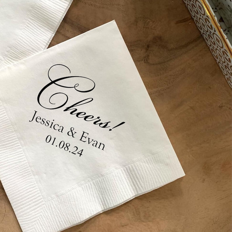 May include: White paper napkin with black text that reads "Cheers! Jessica & Evan 01.08.24".