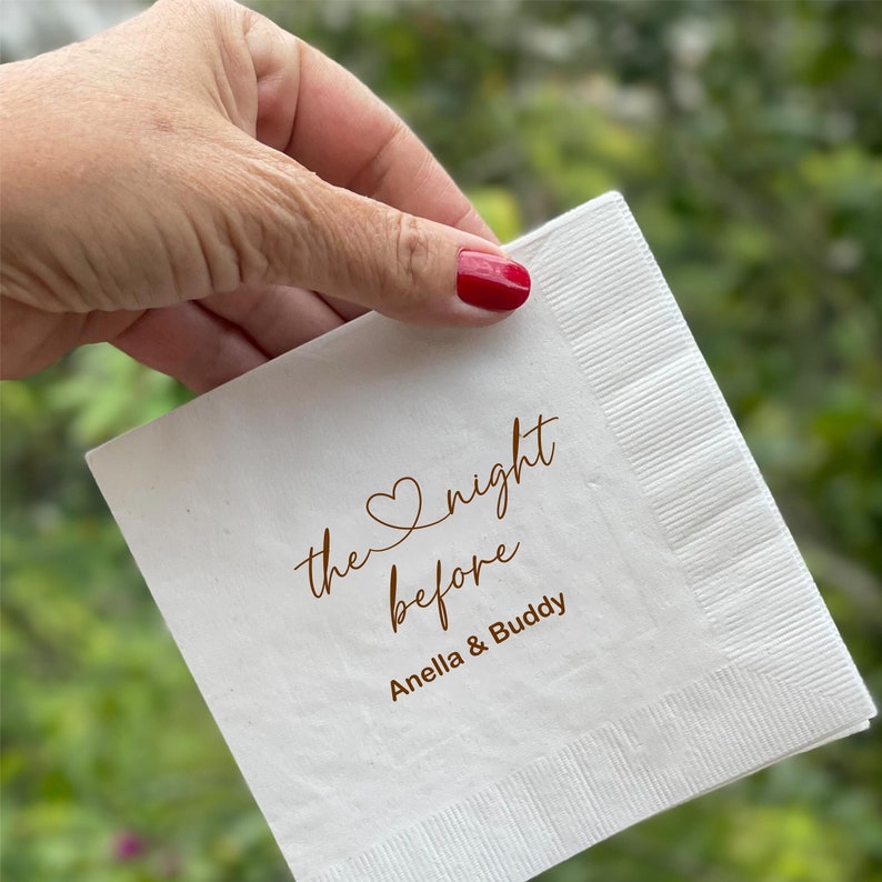 May include: White paper napkin with the text "the night before" in brown cursive script with a heart symbol above the word "night". The text "Anella & Buddy" is printed below in brown cursive script.
