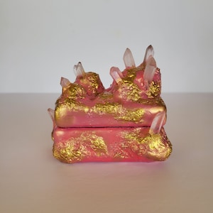 May include: A pink and gold decorative box with a crystal-like design. The box has a hinged lid and is decorated with gold accents and clear crystals.