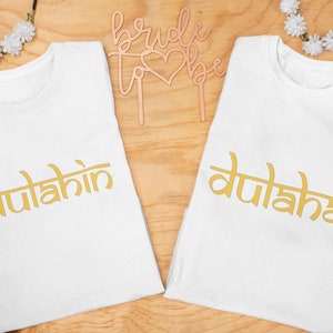 May include: Two white t-shirts with gold text that reads "dulahin" and "dulaha". A rose gold "bride to be" cake topper and floral accents are on a wooden surface. Perfect for a wedding or bridal shower.