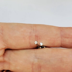 Puede incluir: Un par de pequeños pendientes de botón plateados, cada uno con una sola gema redonda y transparente. Los pendientes se muestran sobre un fondo claro, resaltando su delicado diseño y brillo. Los pendientes se sostienen en una mano.