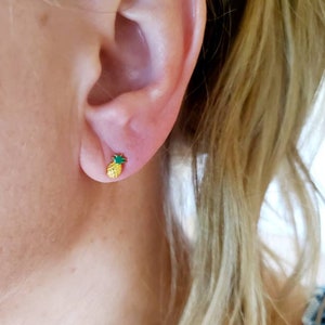 May include: A close-up of a person's ear with a gold-colored pineapple stud earring. The pineapple has a textured yellow body and a green top. The earring is small and sits on the earlobe.
