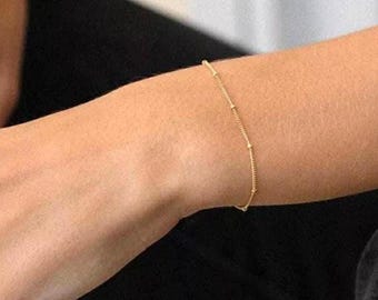 May include: A delicate gold bracelet with small, evenly spaced beads. The bracelet is thin and elegant, designed to be worn on the wrist. The jewelry is simple and minimalist, suitable for everyday wear.