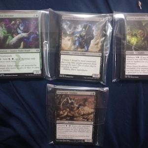 May include: Four Magic: The Gathering trading cards individually sealed in clear plastic sleeves. The cards are Sultai Devotee, Lazotep Behemoth, Grave Scrabbler, and Dregscape Zombie, each featuring unique character illustrations and game text.