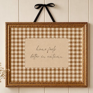 May include: A rectangular wall hanging featuring a gold frame, a brown and white gingham fabric background, and a central beige fabric patch with frayed edges. The text "home feels better in autumn" is printed in black script on the patch.