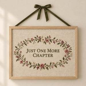 May include: A rectangular wooden frame containing a beige linen fabric with an embroidered floral wreath in shades of pink, red, and green. The text "JUST ONE MORE CHAPTER" is embroidered in the center. An olive green ribbon bow hangs above.