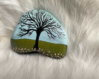May include: Hand-painted rock featuring a black tree silhouette against a light blue sky. The base of the rock has a green field with small pink flower accents. The rock is set against a white, fluffy background.