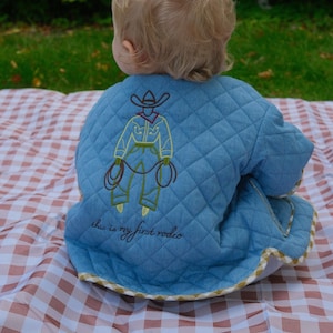 May include: A light blue quilted jacket featuring a yellow and brown embroidered cowboy graphic on the back. Below the embroidery, the text "this is my first rodeo" is stitched in dark thread. The jacket has a tan and white chequered trim.