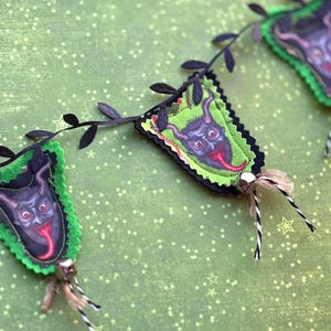 May include: A decorative garland featuring triangular fabric pennants with a dark horned creature graphic, a bright green border, and a black scalloped edge. The pennants are connected by a black vine-like cord with small silver bells and twine.
