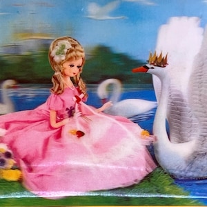 May include: A vintage doll in a pink dress sits near a swan wearing a crown. The doll has blonde hair and is holding a small bouquet. The background features a lake, other swans, and a castle.