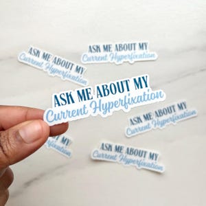 May include: White stickers with the text "ASK ME ABOUT MY Current Hyperfixation" in blue script. Several stickers are scattered on a white surface, with one held by a hand.