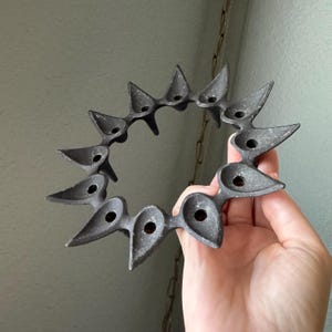 May include: A dark gray metal candle holder with a circular design. The holder features twelve individual candle holders, each with a pointed, leaf-like shape. The overall design is symmetrical and has a rustic, vintage aesthetic.