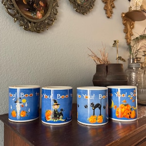 May include: Four ceramic mugs with a blue background featuring a repeating Halloween theme. Each mug displays the text 'Boo to You!' along with illustrations of a ghost, a witch, a black cat, and pumpkins, all accented with small orange pumpkins.