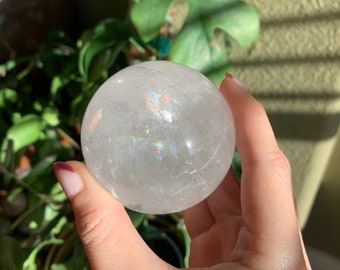 May include: A clear quartz crystal sphere with rainbow iridescence. The sphere is being held in a hand.