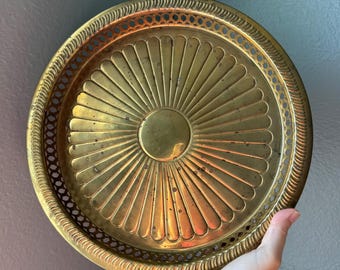 May include: A round brass tray featuring a central circular medallion surrounded by radiating fluted panels. The outer edge has a decorative pierced lattice border with a rope-style rim. The metal surface shows a warm, polished gold-toned finish.