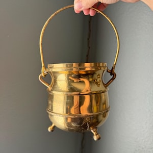 May include: A polished brass pot with a rounded body, a wide rim, and three small feet at the base. The pot features two side handles attached to a single, tall, arched swing handle that extends upward, creating a classic bucket-like silhouette.