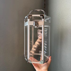 May include: A tall, hexagonal, transparent carrying case with a silver-toned metal frame, reinforced corner caps, and a top handle. The container features a central metal latch mechanism on the front panel, allowing for secure closure of the clear structure.