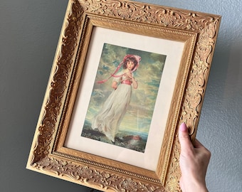 May include: A framed portrait of a young girl in a white dress and pink hat, standing in a field with a pink ribbon blowing in the wind. The frame is ornate and gold.