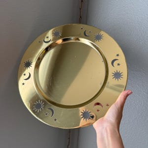 May include: A circular gold-toned metal tray featuring a polished reflective surface. The wide rim is decorated with cut-out shapes of suns, crescent moons, and stars arranged in a repeating pattern around the center.