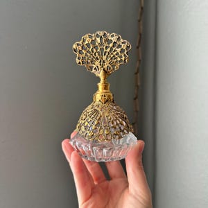 May include: A vintage perfume bottle with a clear glass base and a decorative gold-colored filigree top. The bottle has a detailed, ornate design, and the top features a fan-like shape. The bottle is held in a hand.