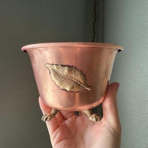 May include: A copper-toned metal bowl featuring a prominent embossed brass leaf design on the front. The bowl is supported by three small, detailed brass feet shaped like animal paws and includes a metal chain attached to the top rim.