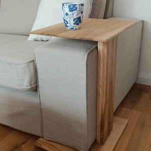 Könnte beinhalten: Ein Beistelltisch aus hellem Holz steht neben einem beigen Sofa. Auf dem Tisch steht eine blau-weiß gemusterte Tasse. Der Tisch ist so konzipiert, dass er über die Armlehne des Sofas passt.