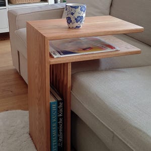 Könnte beinhalten: Beistelltisch aus hellem Holz mit zwei Ablagen und einem vertikalen Zeitschriftenständer. Auf der oberen Ablage steht eine Tasse mit blauem Blumenmuster. Auf den unteren Ablagen liegen eine Zeitschrift und Bücher.