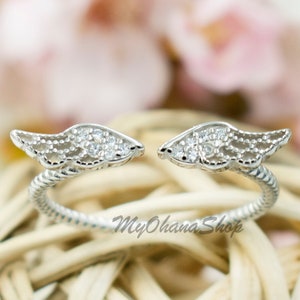 Op de afbeelding: Een zilverkleurige open ring met vleugelvormige uiteinden, elk versierd met kleine, heldere edelstenen. De ring heeft een gedraaid bandontwerp. De achtergrond heeft zachte roze en witte bloemen en een geweven mand.